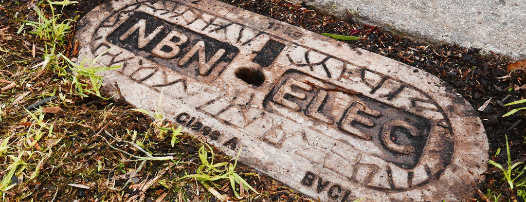 Weathered utility cover embedded in the ground with "NBN ELEC" text.