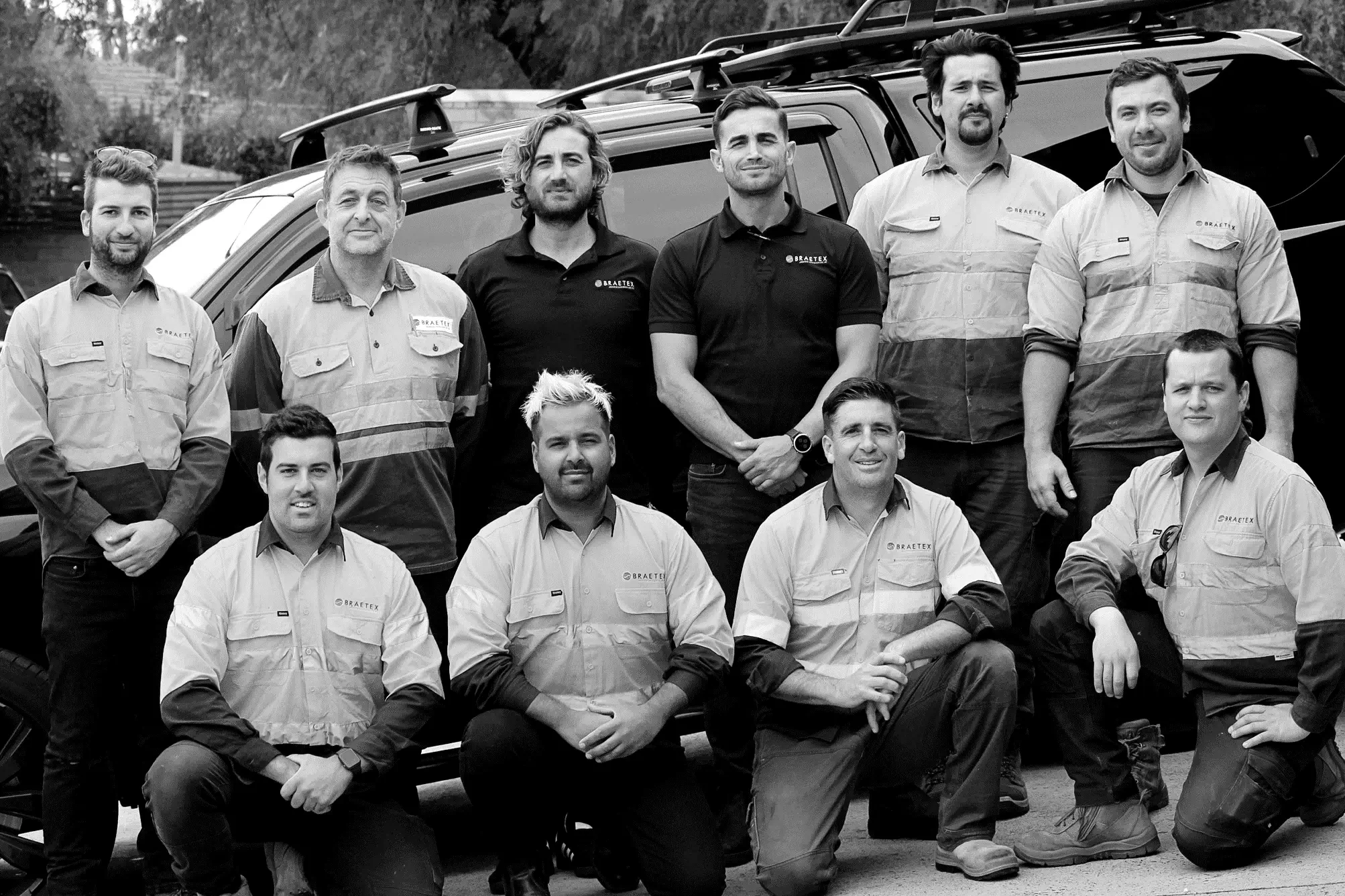 Black and white photo of a professional team of ten male electricians and technicians in work uniforms, standing and kneeling in front of a dark vehicle.